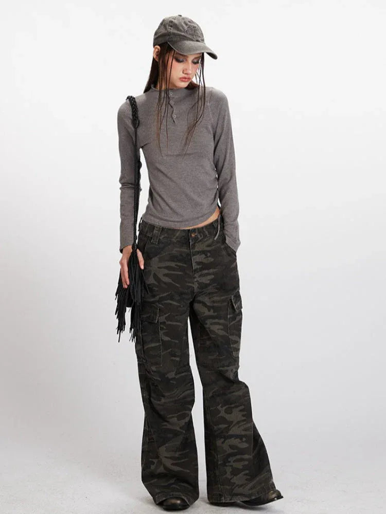 Full-body shot of a model wearing a casual outfit consisting of a grey long-sleeve mock neck cropped top, camouflage cargo pants, a grey baseball cap, and a black fringed crossbody bag.