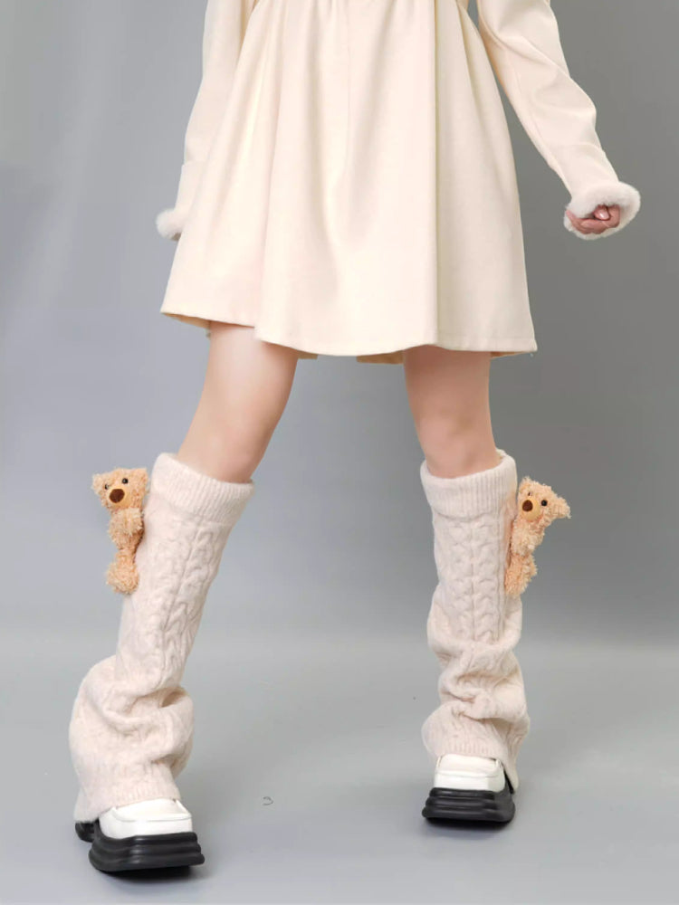 Cream cable knit leg warmers with teddy bear embellishments and pom-poms, worn with a cream dress and tan boots.