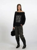 Front view of a woman in a black off-the-shoulder knit sweater with faux fur trim and a graphic print, styled with camouflage cargo pants.