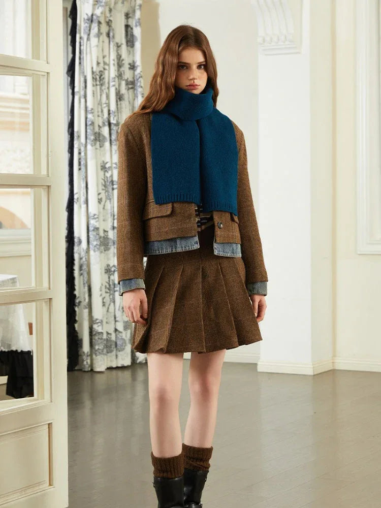 Woman wearing a brown tweed jacket, denim trim, blue knit scarf, and brown pleated skirt.