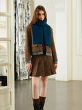 Woman wearing a brown tweed jacket, denim trim, blue knit scarf, and brown pleated skirt.