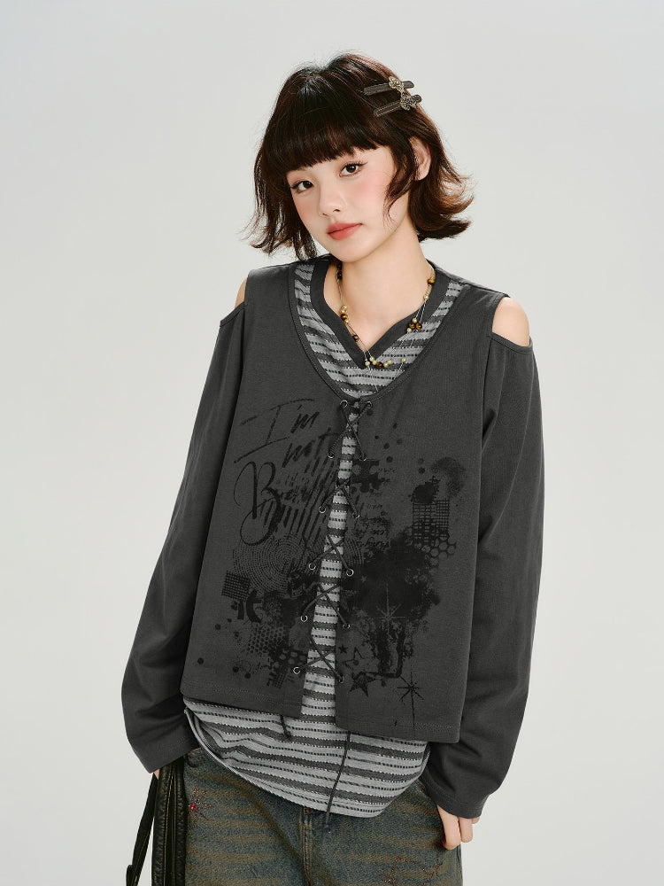 Woman wearing a dark gray graphic print cropped long-sleeve top with cold shoulder cutouts and a lace-up front over a gray and white striped t-shirt.