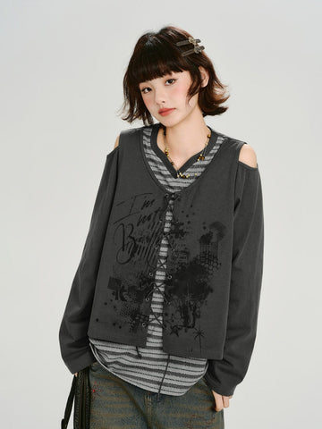 Woman wearing a dark gray graphic print cropped long-sleeve top with cold shoulder cutouts and a lace-up front over a gray and white striped t-shirt.