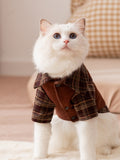 A white cat wearing a brown plaid pet shirt with a collar and button closure.