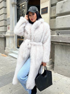 Woman wearing a long, white faux fur coat with a belted waist and lapel collar, holding a black tote bag and wearing a baseball cap.