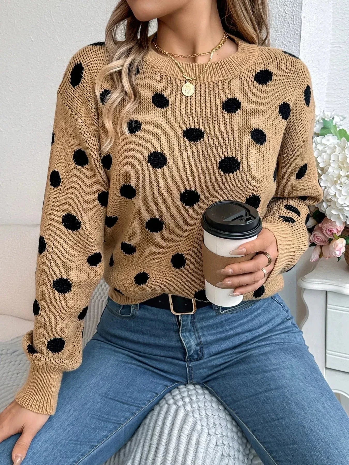 Woman holding a coffee cup while wearing a tan polka dot sweater and blue jeans.