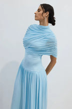 Side profile of a woman wearing a light blue draped jersey maxi dress with a high neckline.