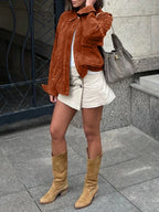 Full body shot of a woman in a brown suede-like bomber jacket, white t-shirt, beige skirt, and brown boots.