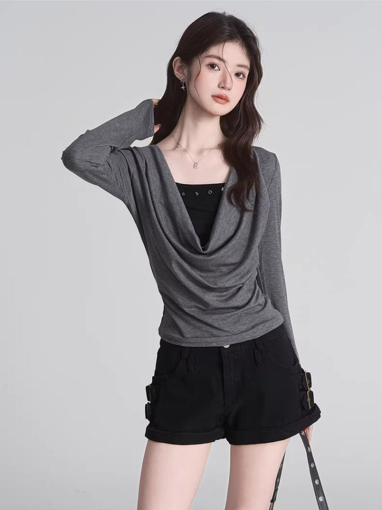 Woman in a grey long-sleeve draped cowl neck top and black denim shorts, posing with hand on hip.