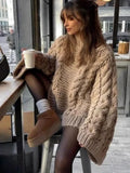 Woman wearing a beige chunky knit oversized sweater with cable knit details, holding a coffee cup.
