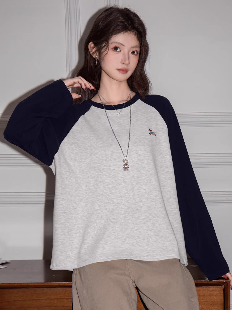 Woman posing with hands on hips wearing a light grey long-sleeve raglan shirt with navy blue sleeves and a small embroidered bird graphic.