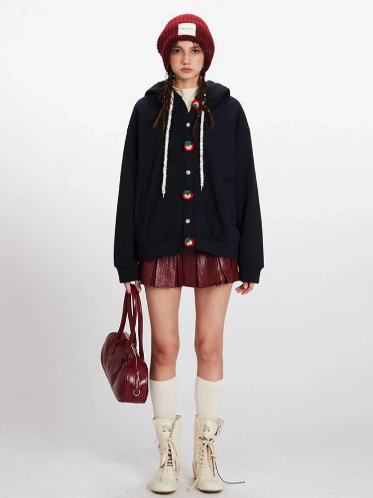 Woman wearing a navy blue hooded fleece jacket with apple-shaped buttons, a red beanie, and a burgundy leather-look pleated skirt. Main image
