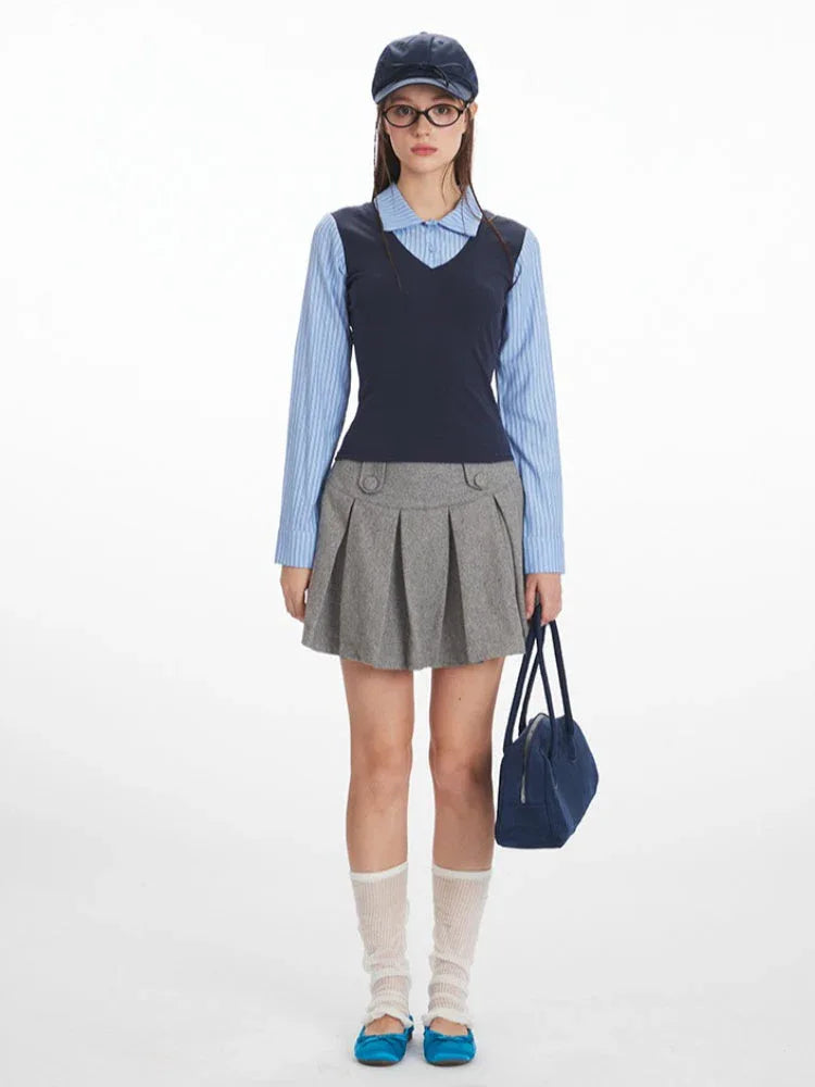 Woman wearing a blue striped collared shirt, navy V-neck vest, and grey pleated skirt, holding a navy handbag.