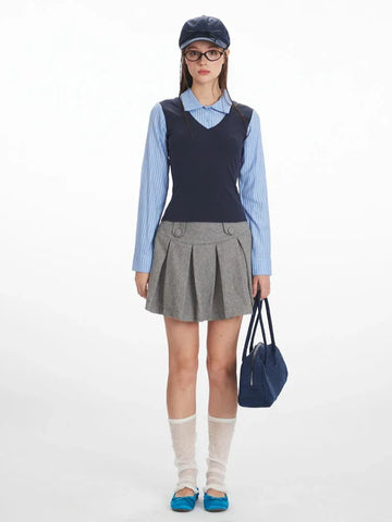 Woman wearing a blue striped collared shirt, navy V-neck vest, and grey pleated skirt, holding a navy handbag.
