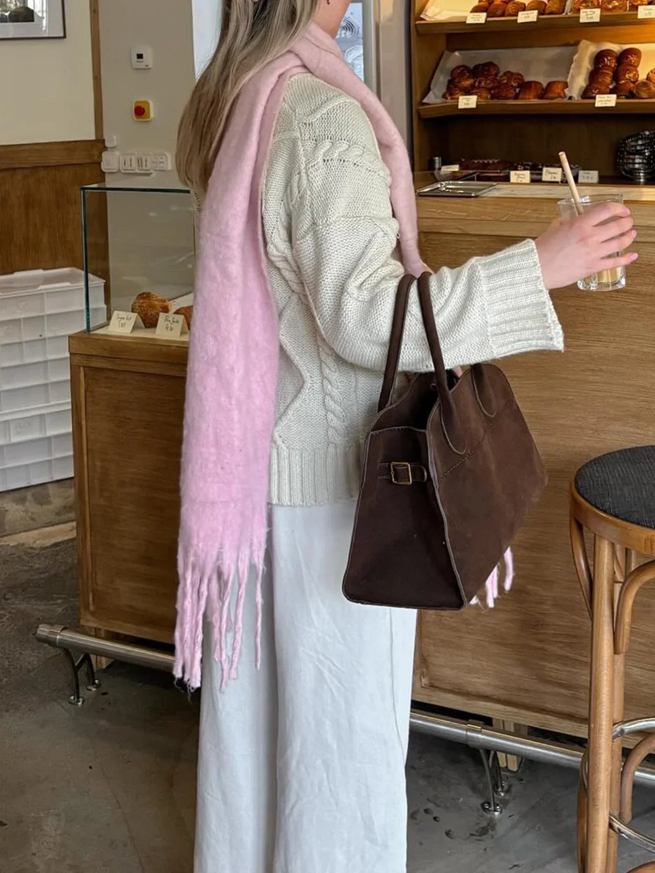 Woman wearing a cream cable knit sweater, white pants, and a pink scarf, carrying a brown handbag.