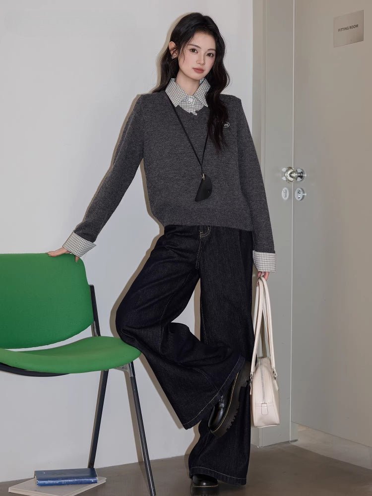 Full body shot of a woman wearing a grey V-neck sweater, checkered shirt, and dark wide-leg jeans