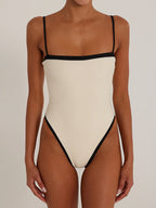 Woman wearing a cream one-piece swimsuit with black contrast trim and spaghetti straps.