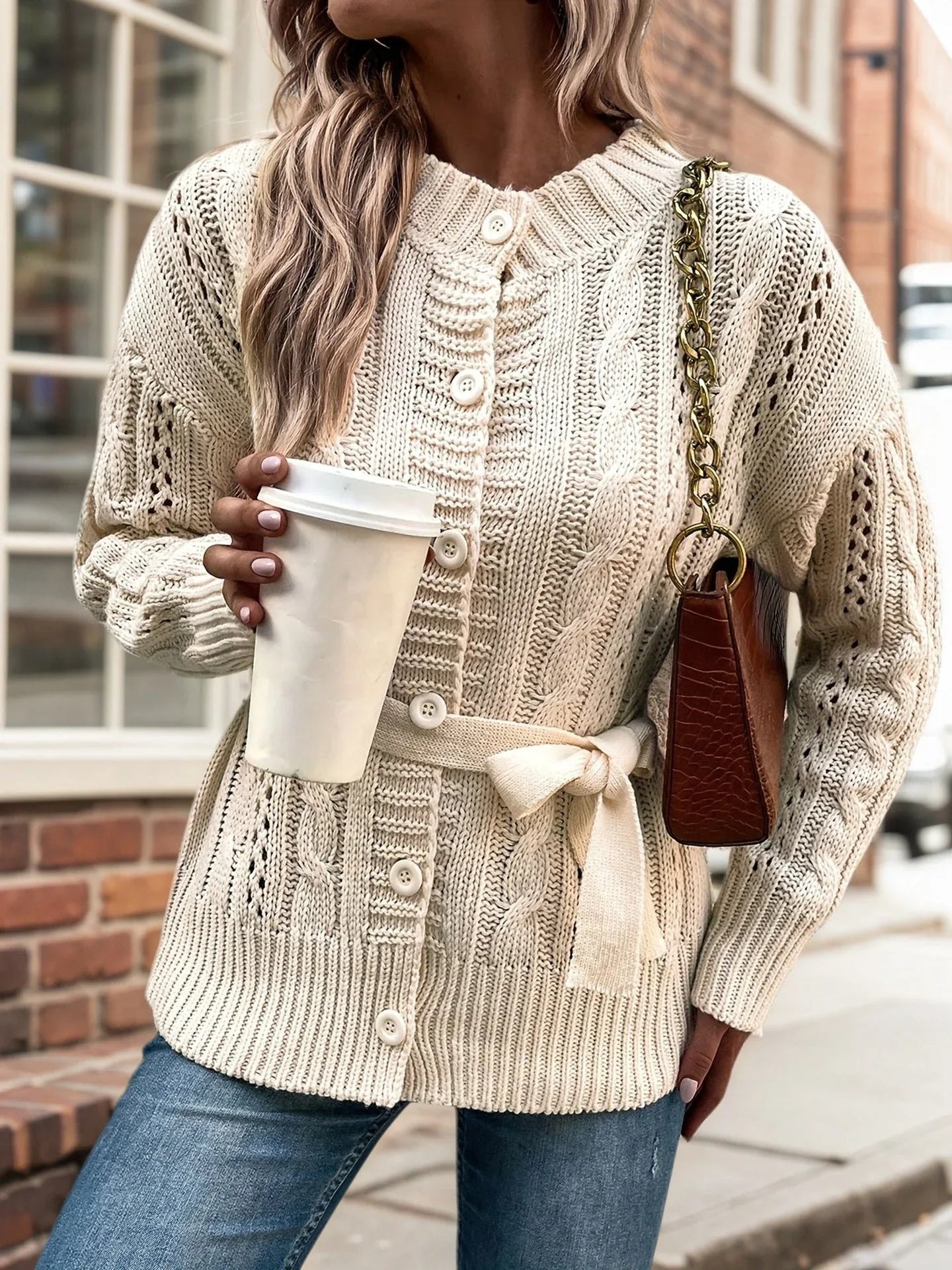 Woman in a cream knit cardigan with a belt tied at the waist, holding a coffee cup and a handbag.