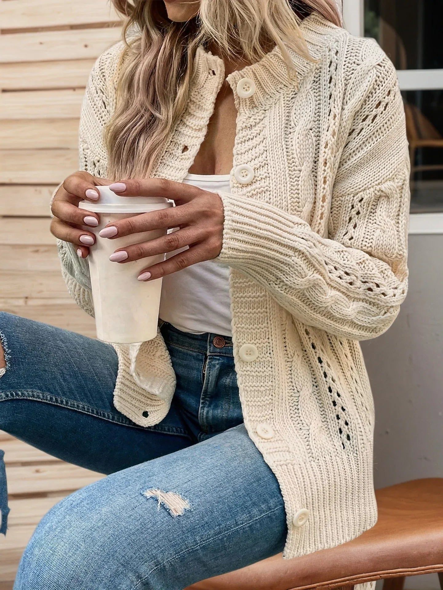 Woman wearing a cream cable knit cardigan with openwork details, holding a coffee cup.