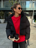 Woman holding two coffee cups while wearing a black hooded puffer jacket with a red t-shirt and black pants.