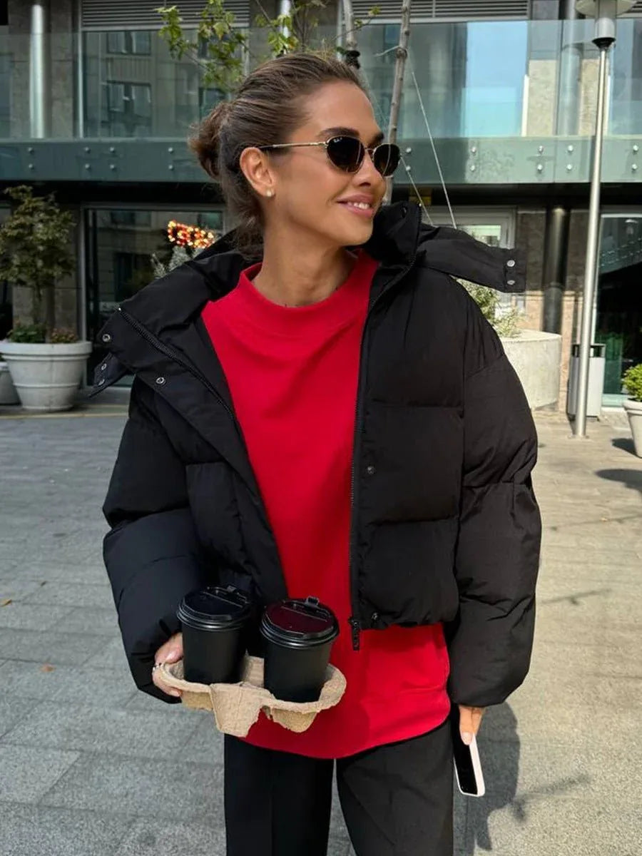 Woman holding two coffee cups while wearing a black hooded puffer jacket with a red t-shirt and black pants.