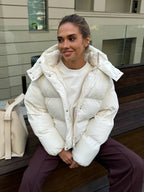 Woman sitting on a bench wearing a white hooded puffer jacket with a white t-shirt and brown pants.