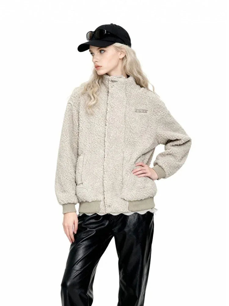 Woman posing in a beige sherpa fleece bomber jacket with a high collar, ribbed cuffs and hem, and an embroidered logo. She is wearing black leather pants and a black baseball cap with sunglasses.