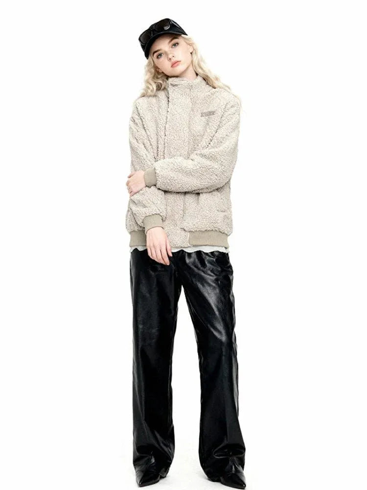 Woman wearing a beige sherpa fleece bomber jacket with a high collar, ribbed cuffs and hem, and an embroidered logo. She is also wearing black leather pants and a black baseball cap.