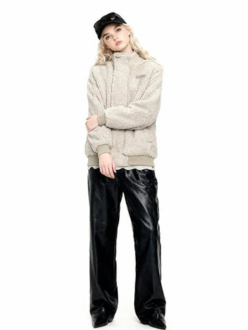 Woman wearing a beige sherpa fleece bomber jacket with a high collar, ribbed cuffs and hem, and an embroidered logo. She is also wearing black leather pants and a black baseball cap.