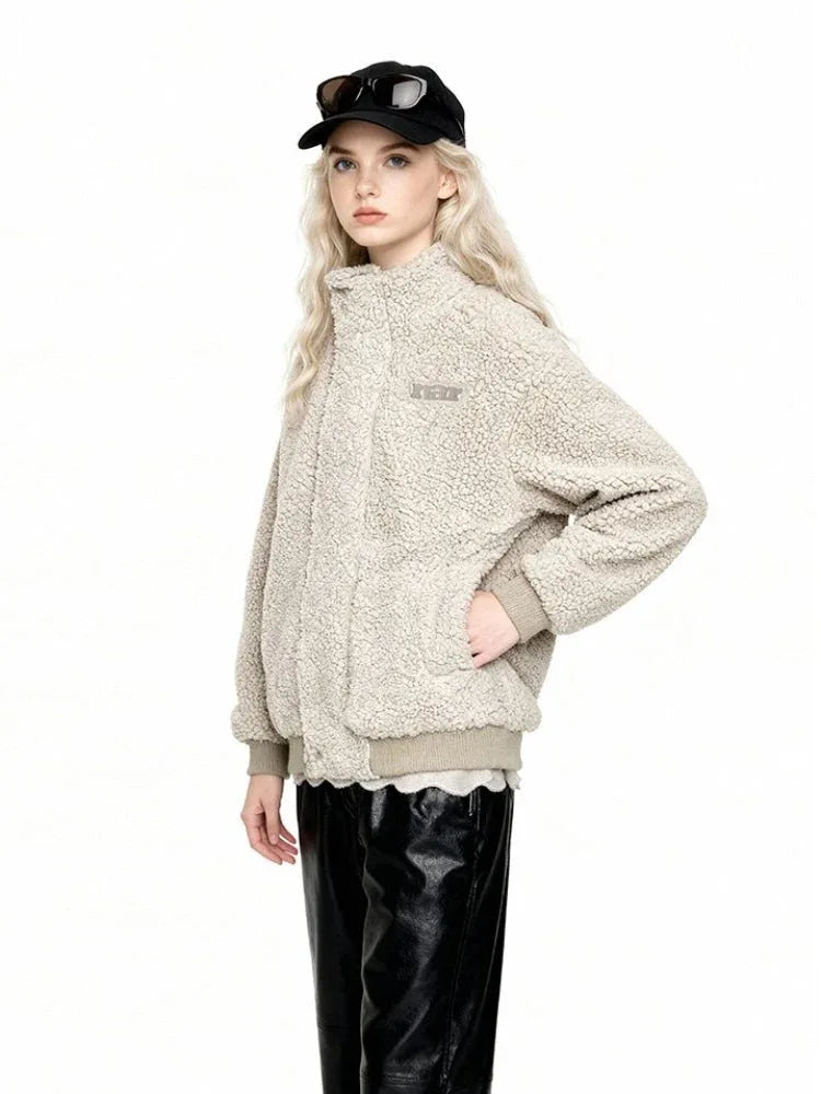 Three-quarter view of a woman wearing a beige sherpa fleece bomber jacket with a high collar, ribbed cuffs and hem, and an embroidered logo. She is wearing black leather pants and a black baseball cap with sunglasses.