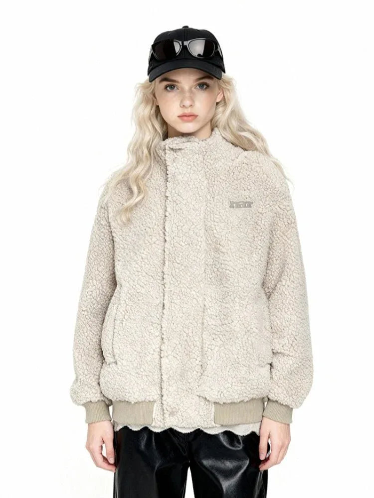 Front view of a woman wearing a beige sherpa fleece bomber jacket with a high collar, ribbed cuffs and hem, and an embroidered logo. She is wearing black leather pants and a black baseball cap with sunglasses.