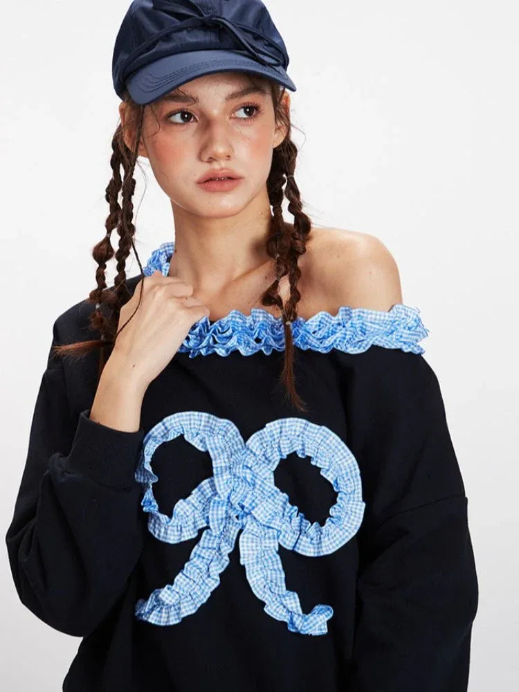 Close-up of the neckline and graphic detail on a black off-the-shoulder sweatshirt with blue gingham ruffled trim and a textured blue bow appliqué.