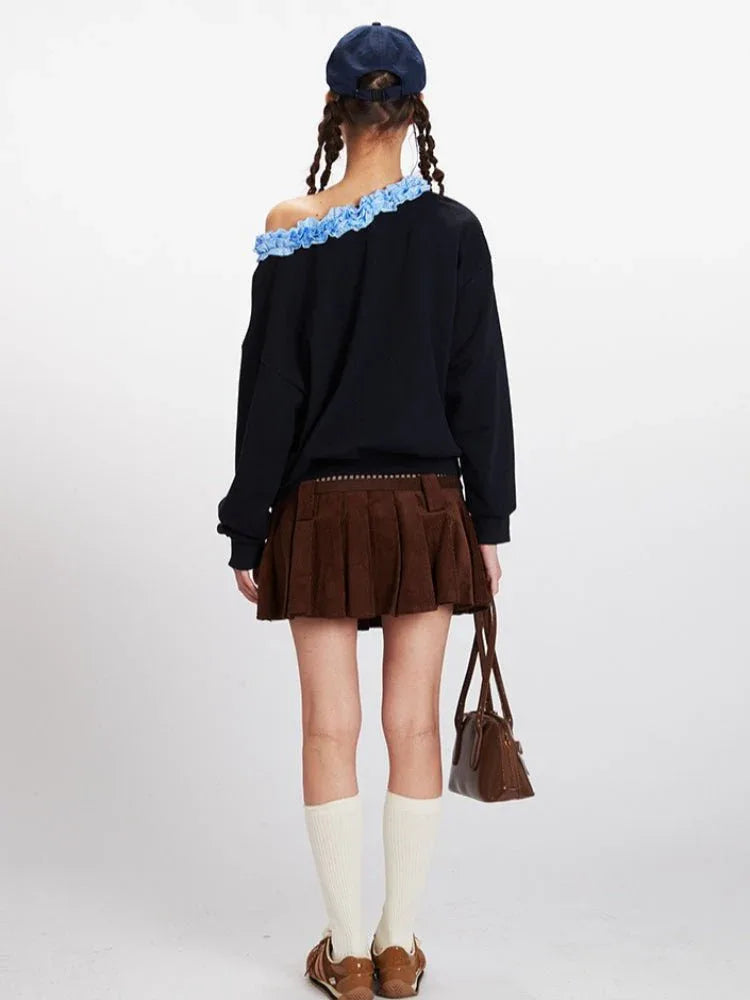 Woman wearing a black long-sleeve sweatshirt with a blue bow graphic and ruffled off-the-shoulder neckline, paired with a brown corduroy pleated skirt and white socks.