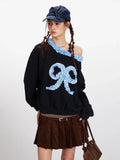 Model posing in a black off-the-shoulder sweatshirt featuring a large blue gingham bow appliqué and ruffled trim.