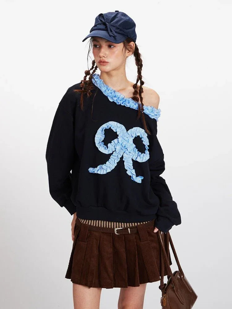 Model posing in a black off-the-shoulder sweatshirt featuring a large blue gingham bow appliqué and ruffled trim.