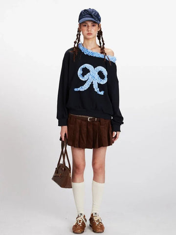 Woman wearing a black long-sleeve sweatshirt with a blue bow graphic and ruffled off-the-shoulder neckline, paired with a brown corduroy pleated skirt and white socks.