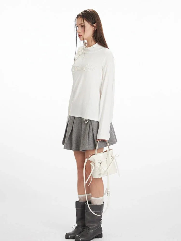 Woman wearing a white long-sleeve top with an asymmetrical shoulder cut-out and a bow detail, paired with a grey pleated skirt and black boots.