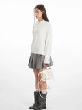 Side view of a woman in a white long-sleeve top with a mock neck and cut-out shoulder, styled with a grey pleated skirt and boots.