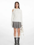 Woman wearing a white long-sleeve top with an asymmetrical shoulder cut-out and a bow detail, paired with a grey pleated skirt and black boots.