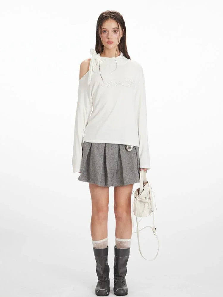 Woman wearing a white long-sleeve top with an asymmetrical shoulder cut-out and a bow detail, paired with a grey pleated skirt and black boots.