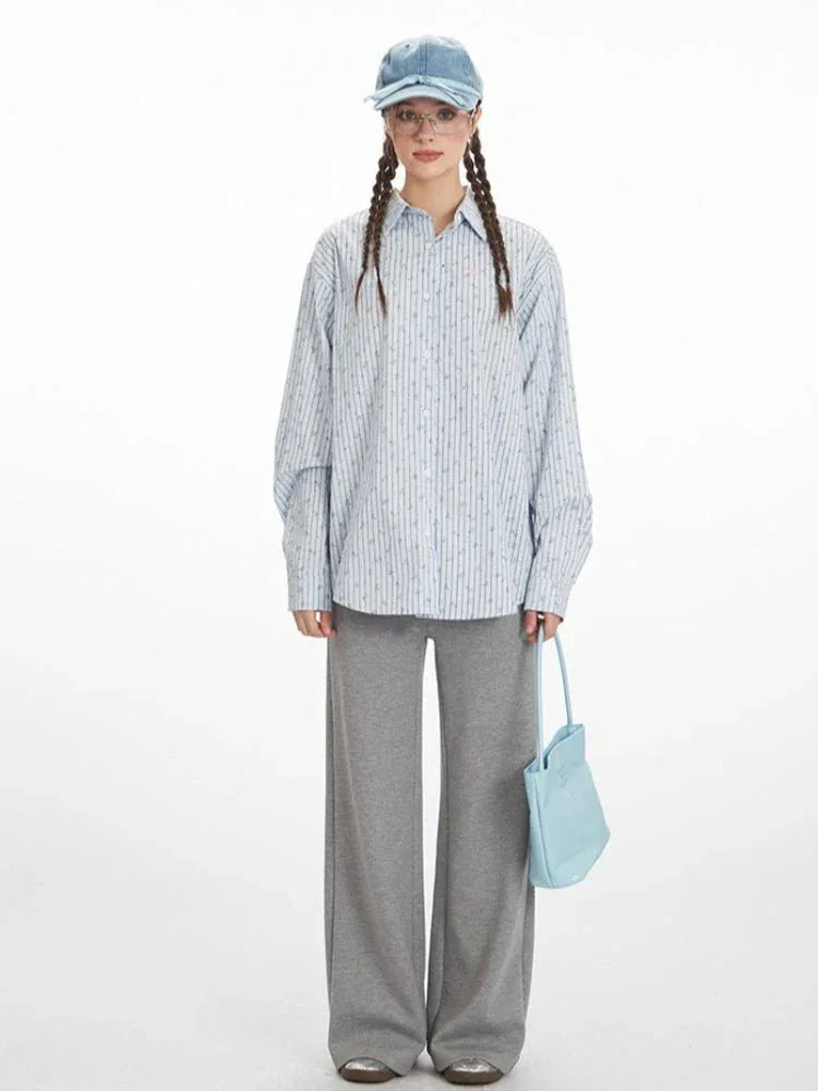 Woman wearing a light blue pinstripe button-down shirt with an open back tied with strings, paired with grey wide-leg pants and a light blue cap.