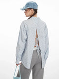 Side view of a woman wearing a light blue and white vertical striped long-sleeve shirt with an open back and tie detail, highlighting the silhouette and back design.