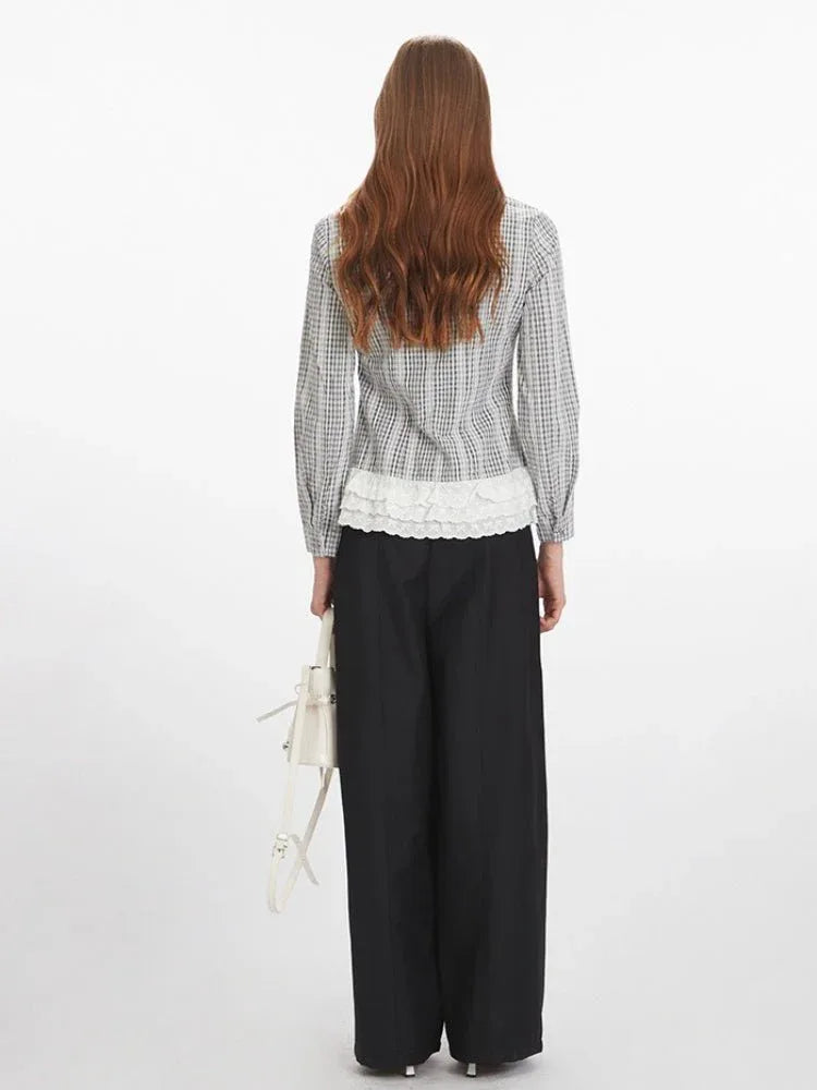 Woman wearing a long-sleeved collared blouse with a plaid pattern and white lace trim at the hem, paired with black wide-leg pants and a white handbag.