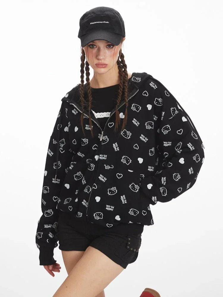 Woman posing in a black zip-up hoodie with a repeating white Hello Kitty and heart graphic print, black shorts, and knee-high socks, with a black baseball cap.