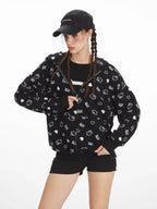 Woman in a black zip-up hoodie with a repeating white Hello Kitty and heart graphic print, black shorts, and knee-high socks, holding a red handbag.