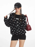 Front view of a woman wearing a black zip-up hoodie with a repeating white Hello Kitty and heart graphic print, black shorts, and a black baseball cap.