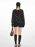 Back view of a woman in a black zip-up hoodie with a repeating white Hello Kitty and heart graphic print, black shorts, and knee-high socks.