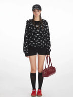 Woman wearing a black zip-up hoodie with a repeating white Hello Kitty and heart graphic print, paired with black shorts, knee-high socks, and red sneakers.