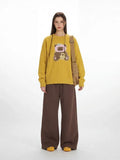 Woman wearing a yellow long-sleeved crew neck sweater with a teddy bear appliqué, paired with brown wide-leg pants and a tan crossbody bag.