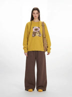Woman wearing a yellow long-sleeved crew neck sweater with a teddy bear appliqué, paired with brown wide-leg pants and a tan crossbody bag.